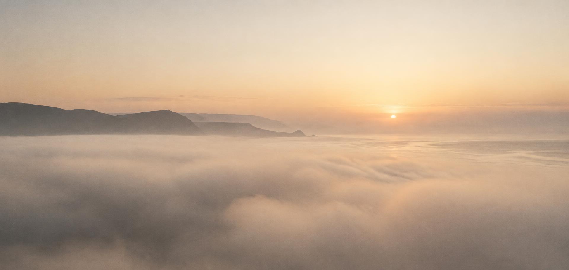 A warm horizon at dawn, a faint coastline emerging through pale fog